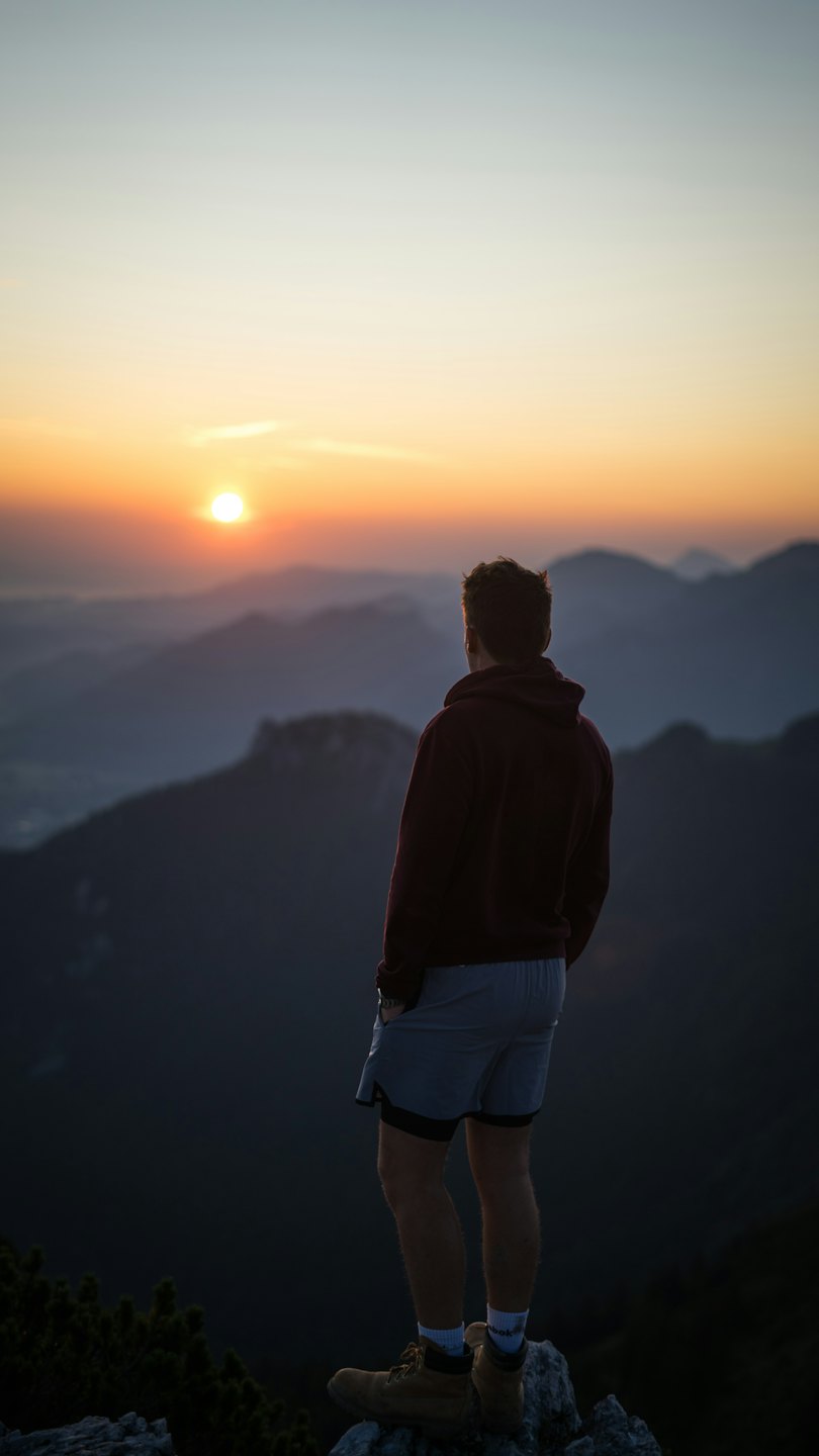 Escursionista in piedi sulla cima rocciosa di una montagna con vista su catene montuose sovrapposte.